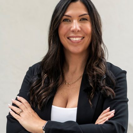 A smiling woman with long, dark hair, wearing a black blazer and a gold chain, with her arms crossed.