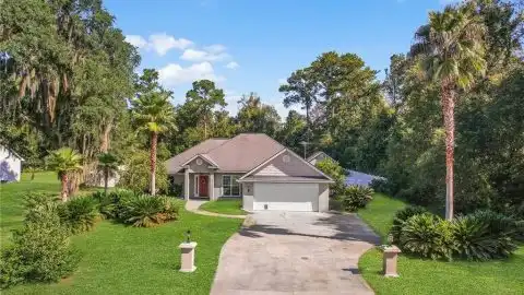 A light-colored single-story house with a gray roof and two-car garage sits on a green, tree-filled lawn at 1155 Sapelo Cir SE, Darien, GA.