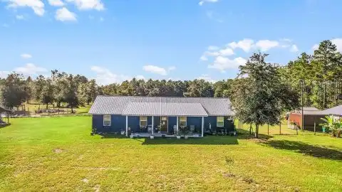A blue ranch-style house with a porch sits in a grassy field under a cloudy blue sky.