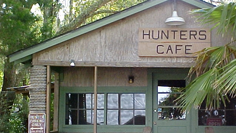 A rustic wooden building with a green roof and a “HUNTERS CAFE” sign stands amid trees and foliage.