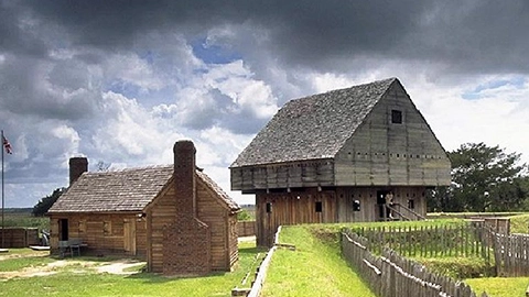 Fort King George with a two-story blockhouse and smaller building under a cloudy sky, surrounded by grass and a wooden fence.